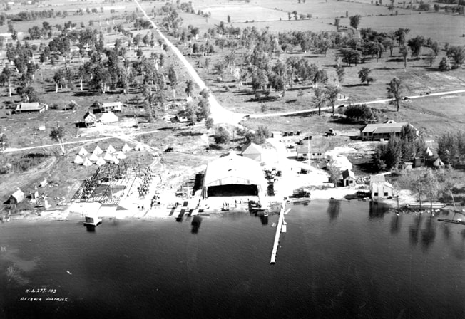Aerial view of Shirleys Bay in mid-1927 looking south along Range Road, which trails away into the distance. The building to the right and above the copse of trees is the officers’ mess and quarters. The tents to the left of the Bessonneau hangar are the accommodations for the airmen. Work has just started on the new hangar.